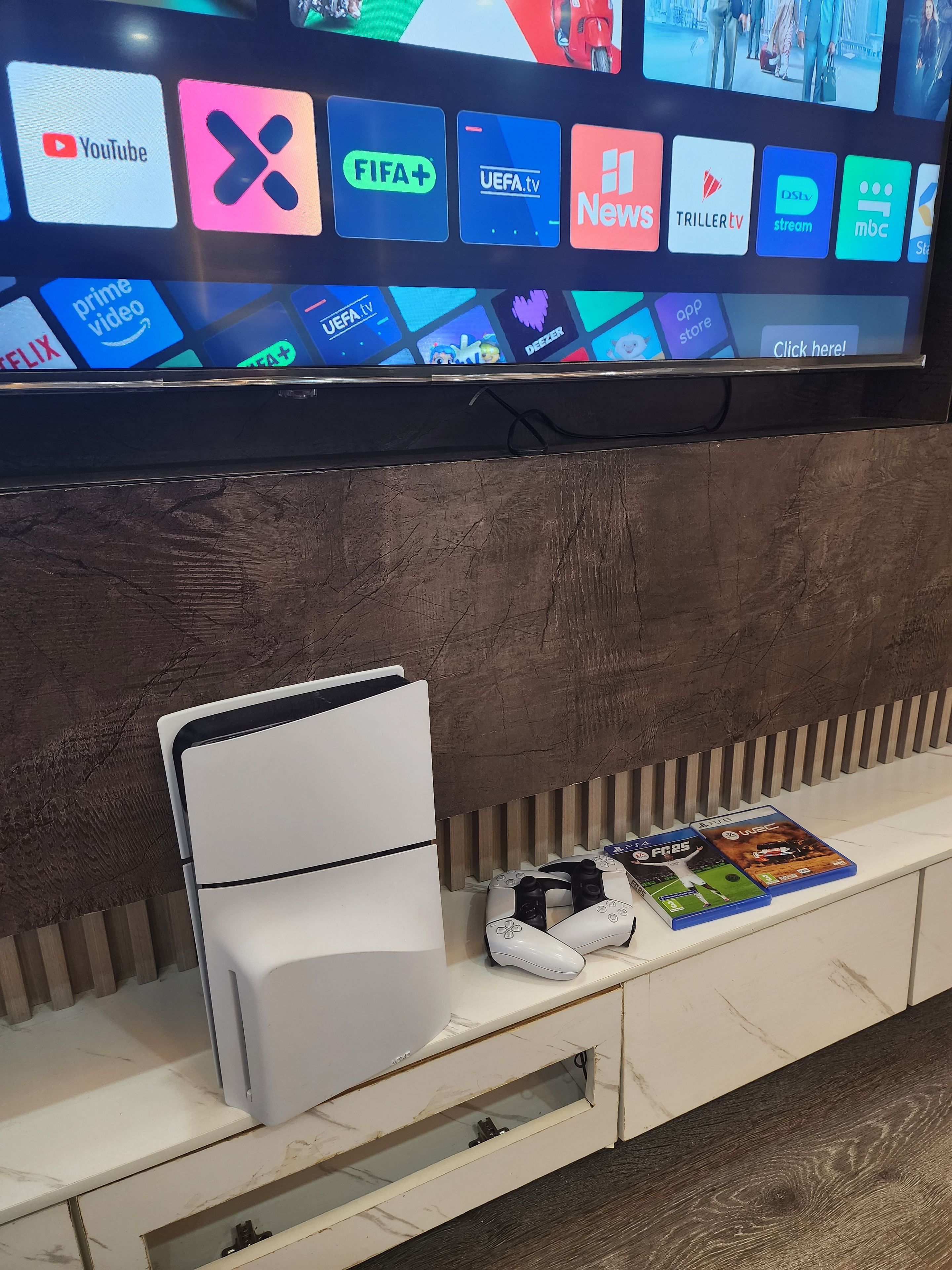 Un televisor de pantalla plana montado en la pared mostrando diversos servicios de transmisión. Debajo del televisor, hay una consola blanca con un control de videojuegos y dos cajas de videojuegos.