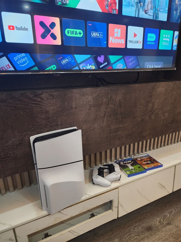 Un televisor de pantalla plana montado en la pared mostrando diversos servicios de transmisión. Debajo del televisor, hay una consola blanca con un control de videojuegos y dos cajas de videojuegos.