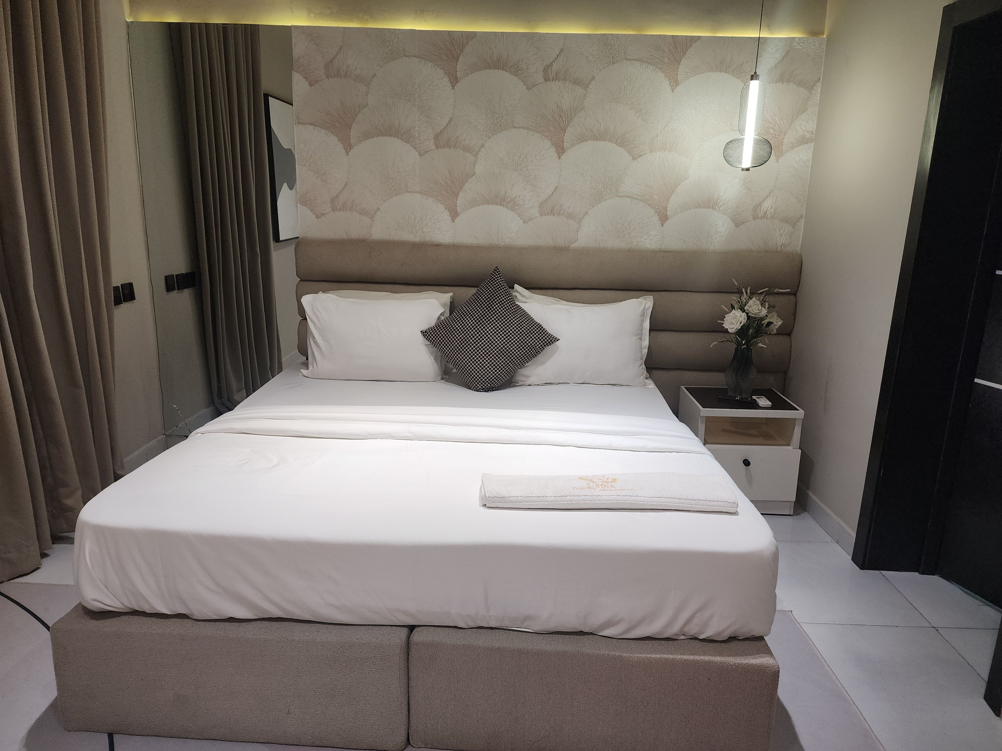 Un lit bien fait, recouvert d’une couvre-lit blanche, agrémenté d’un coussin décoratif et d’une table de chevet sur laquelle est posé un vase de fleurs.