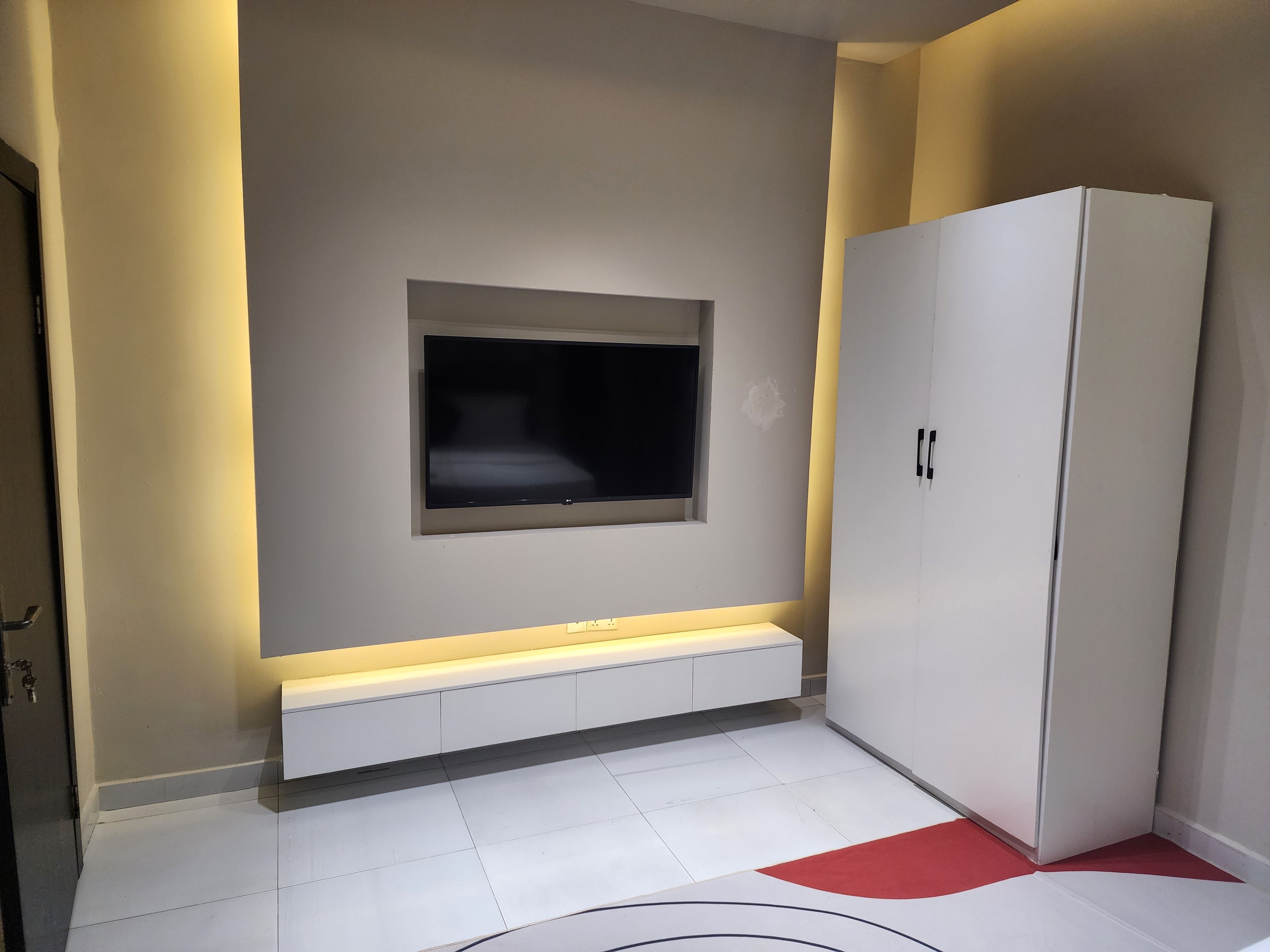 Une pièce dotée d’un téléviseur à écran plat fixé au mur, d’une armoire blanche et d’un tapis rouge qui apporte une touche de couleur au sol.