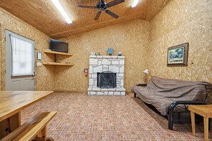 A room with a fireplace, a ceiling fan, a television on a shelf, a bed, and a table. - Riverside Cabins at Cypress Bend #11 @ (Concan)