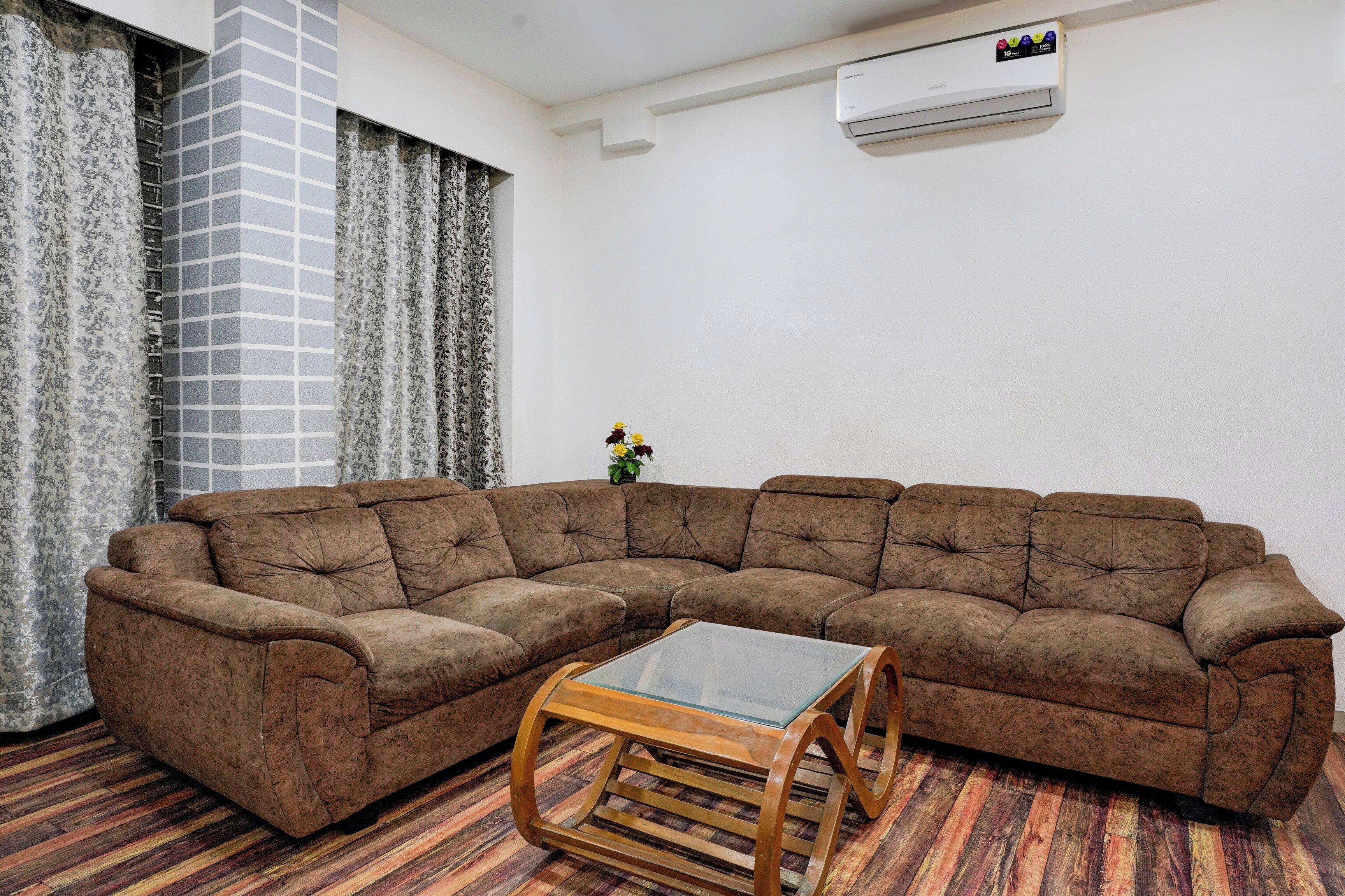 A living room with a sectional sofa, a glass-top coffee table, patterned curtains, and an air conditioning unit on the wall.