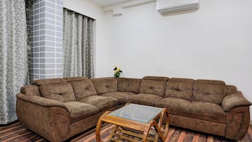 A living room with a sectional sofa, a glass-top coffee table, patterned curtains, and an air conditioning unit on the wall.