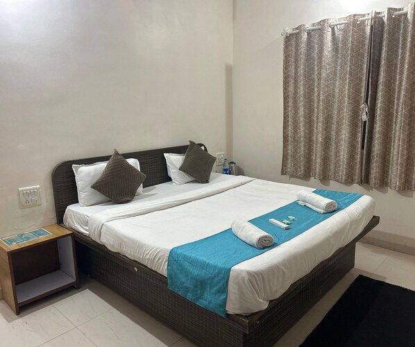 A neatly made bed with pillows, a blue towel, and a small table with a water bottle and a remote control. - Classic Oraa Stay (Mahabaleshwar)