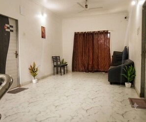 A room with a marble floor, a ceiling fan, a door with a checkered pattern, a brown curtain, a black sofa, and two potted plants. - Classic Oraa Stay (Mahabaleshwar)