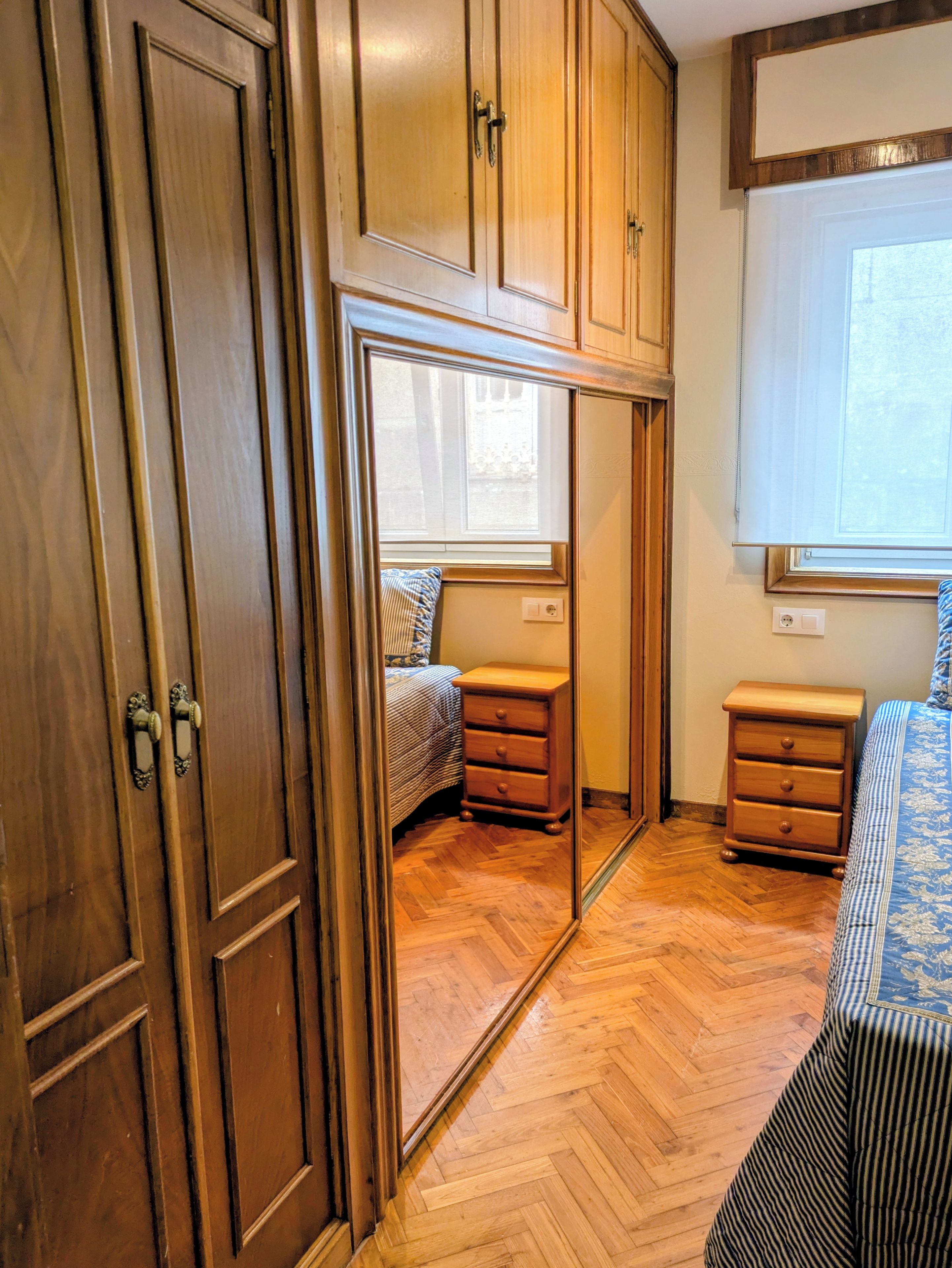 Un dormitorio con muebles de madera, un espejo, una ventana y un suelo con patrón de espiga.