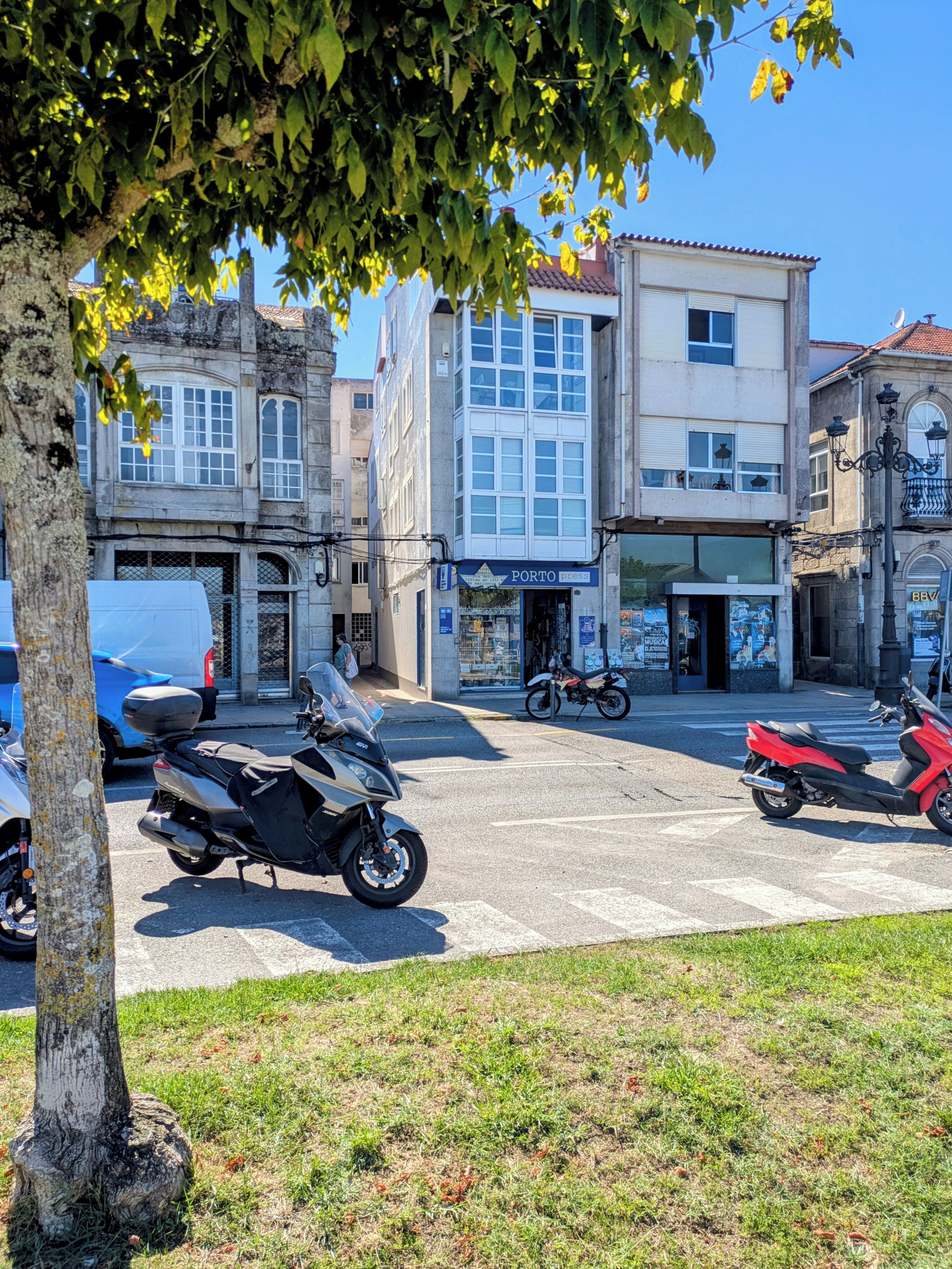 Straßenansicht mit abgestellten Motorrädern, einem Gebäude mit dem Schild 'PORTO press' und einem Baum im Vordergrund.