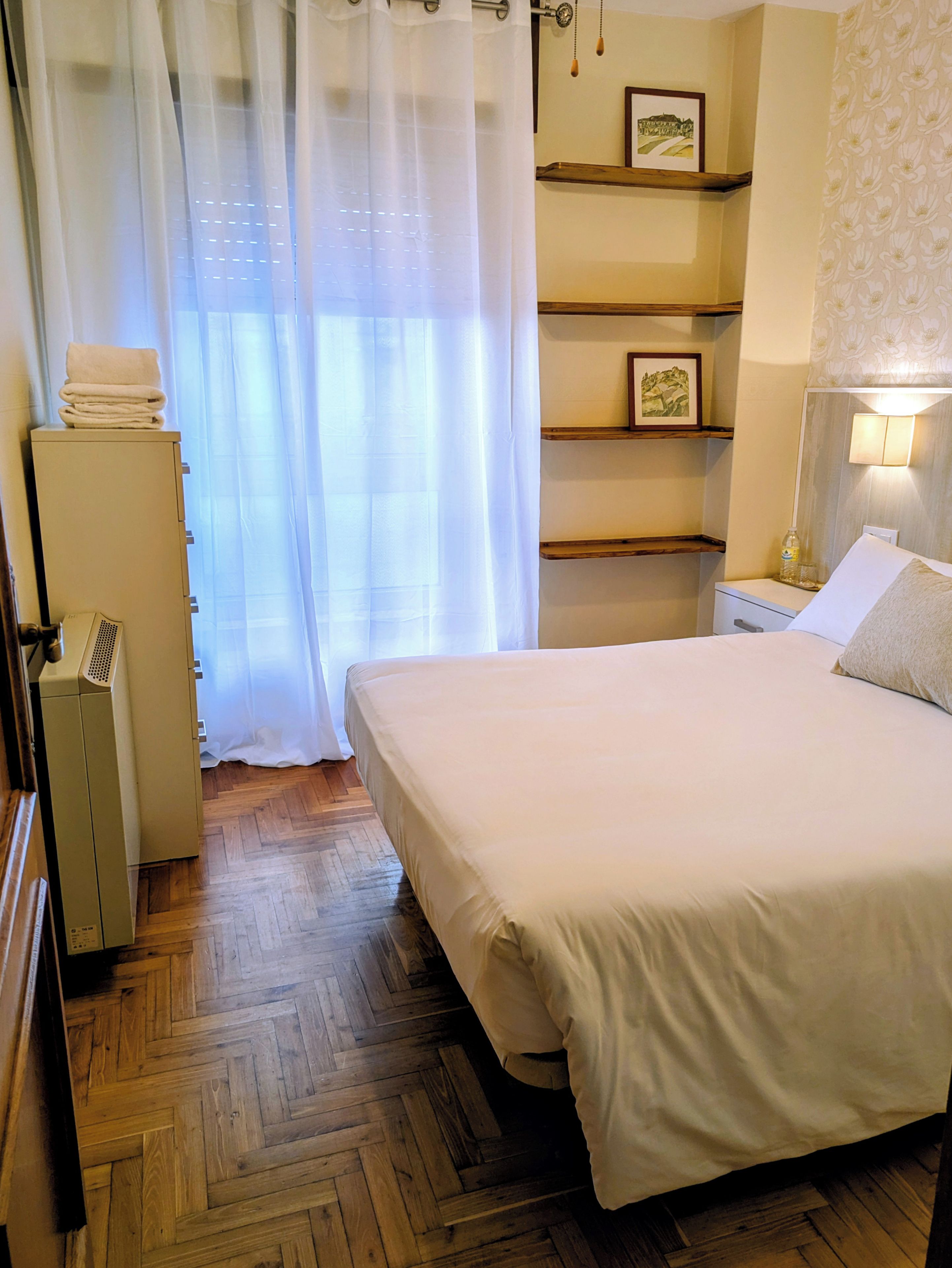 Ein Hotelzimmer mit Bett, Schrank, Nachttisch mit Lampe und einer an der Wand befestigten Ablage mit Bildern.