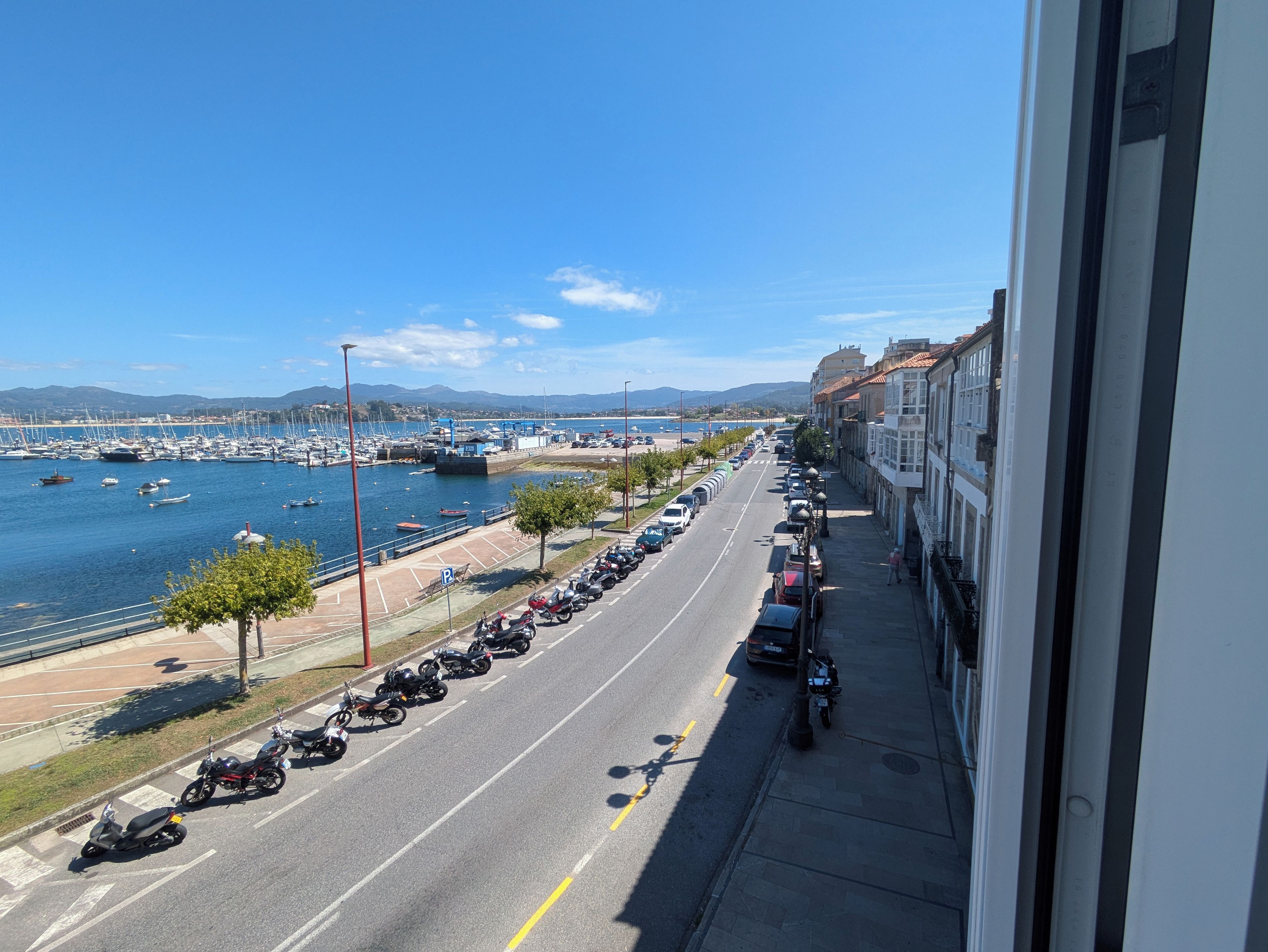 Una carretera costera con motocicletas y automóviles estacionados, un puerto deportivo con barcos y una vista despejada del mar y montañas distantes.