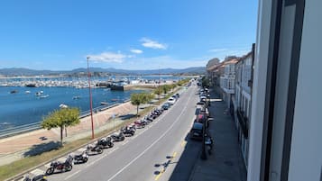 Una carretera costera con motocicletas y automóviles estacionados, un puerto deportivo con barcos y una vista despejada del mar y montañas distantes.
