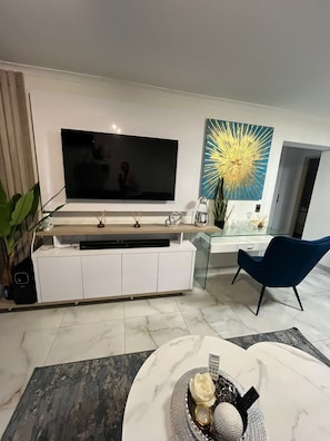 A modern living room with a flat-screen TV, a glass coffee table, a blue chair, and a decorative wall piece. - Stylish Stay with Pools & Gym by Ctha (Cape Town)