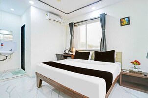 Habitación de hotel con cama, escritorio, silla, ventana con cortinas, mesita de noche con lámpara y aire acondicionado instalado en la pared.
