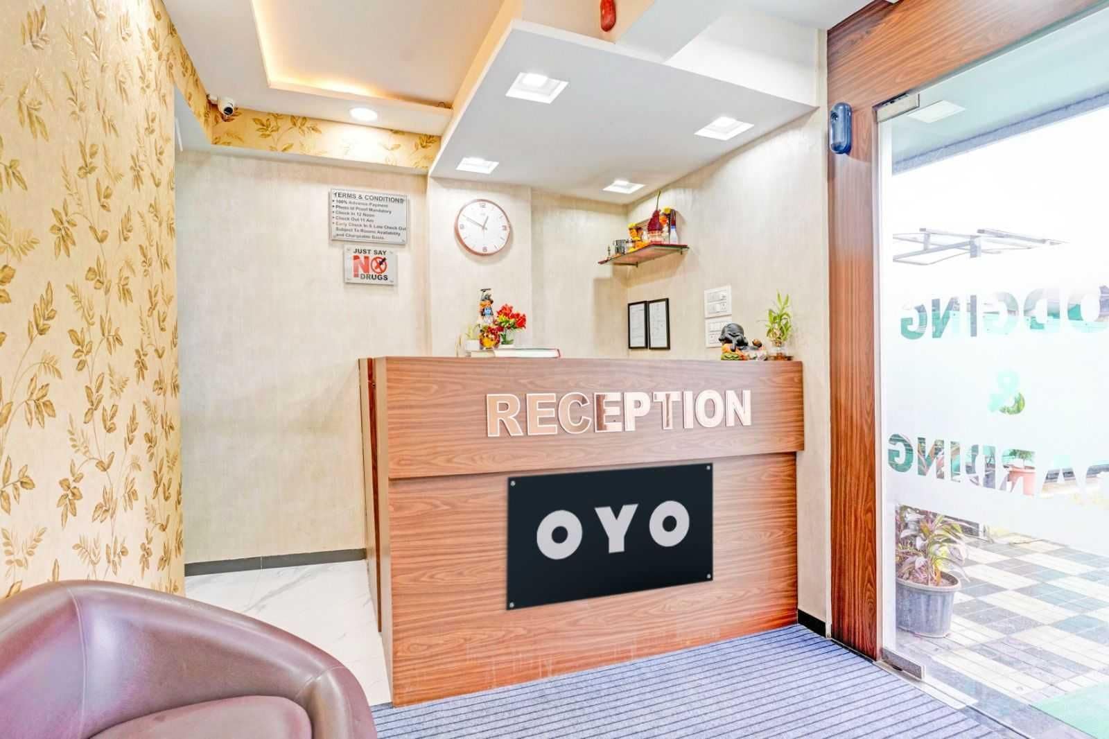 Un comptoir d’accueil avec un revêtement en bois et une enseigne noire sur laquelle on peut lire « RÉCEPTION OYO ». Il y a une horloge au mur, au-dessus du comptoir, et une petite étagère avec divers objets.