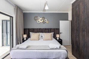 Une chambre moderne avec un grand lit, deux tables de chevet, un luminaire fixé au mur et une vue sur l’extérieur, visible à travers une porte vitrée.