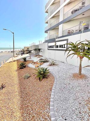 A building with a sign reading 'Sunset on Hill' and a landscaped area with plants and pebbles. - Wonderful 2-bedroom apartment in welcoming Bloubergstrand neighborhood (Cape Town)