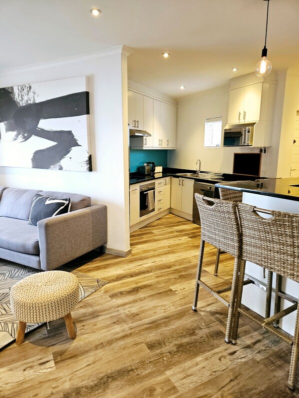 A modern kitchen with white cabinets, a dining area with a bar stool, and a grey sofa. - Wonderful 2-bedroom apartment in welcoming Bloubergstrand neighborhood (Cape Town)