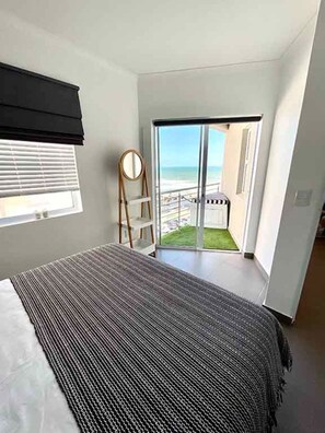 Ein Schlafzimmer mit einem Bett, einem Fenster, einer Tür, die auf einen Balkon führt, und Blick auf den Strand.