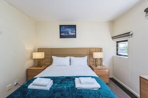 A hotel room with a bed, two bedside tables with lamps, a window with blinds, and a framed picture on the wall. - Chic Queens Gate Duplex by Ctha (Cape Town)