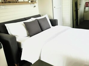 A bed with a white duvet and grey pillows in a room with white walls and a wooden shelf above. - Buzzards Lodge (Bodmin)