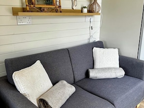 A grey sofa with two white pillows and a rolled-up blanket. A wooden shelf above holds decorative items. - Buzzards Lodge (Bodmin)