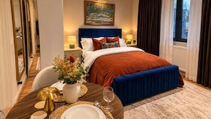 A hotel room with a large bed, a dining table set for two, a vase of flowers, and a painting on the wall. - Kings Cross 1-bed Apt, Sleeps 3 With Great Views (London)