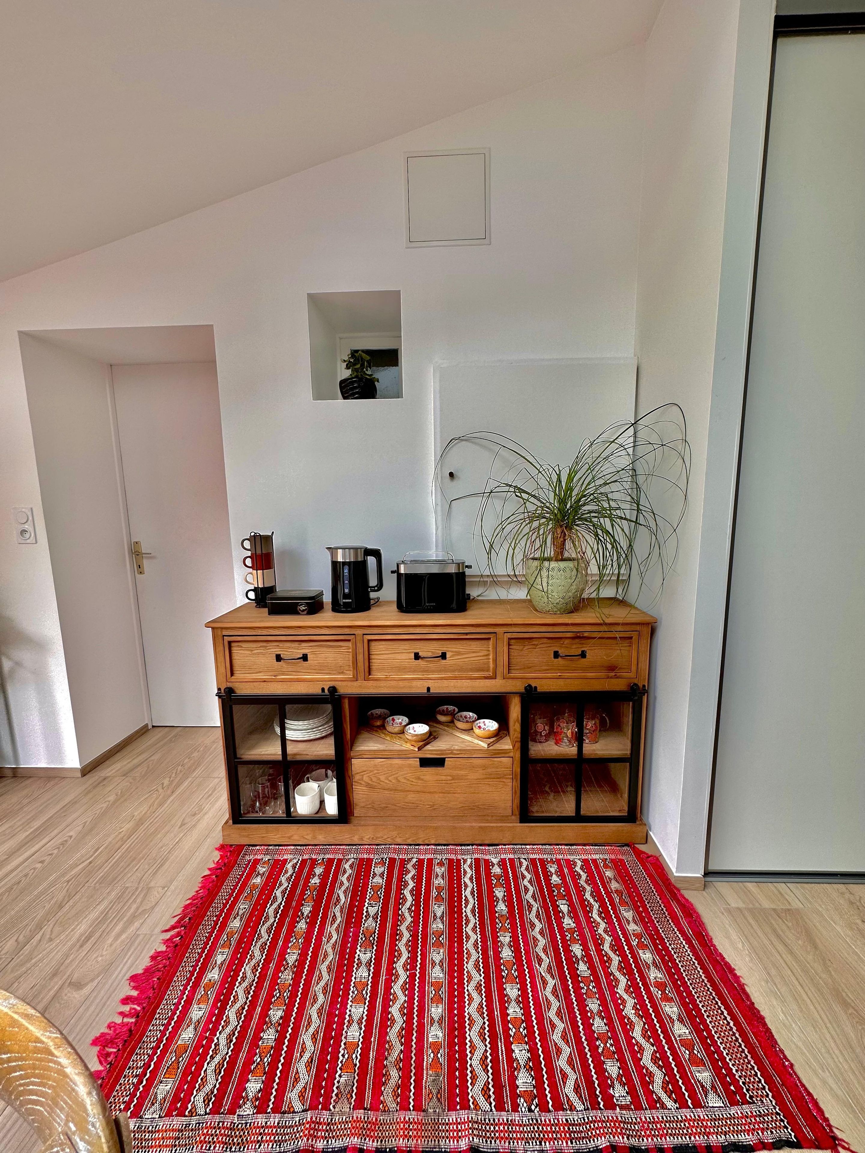 Une pièce avec un meuble en bois, un tapis rouge à motifs et une plante en pot.