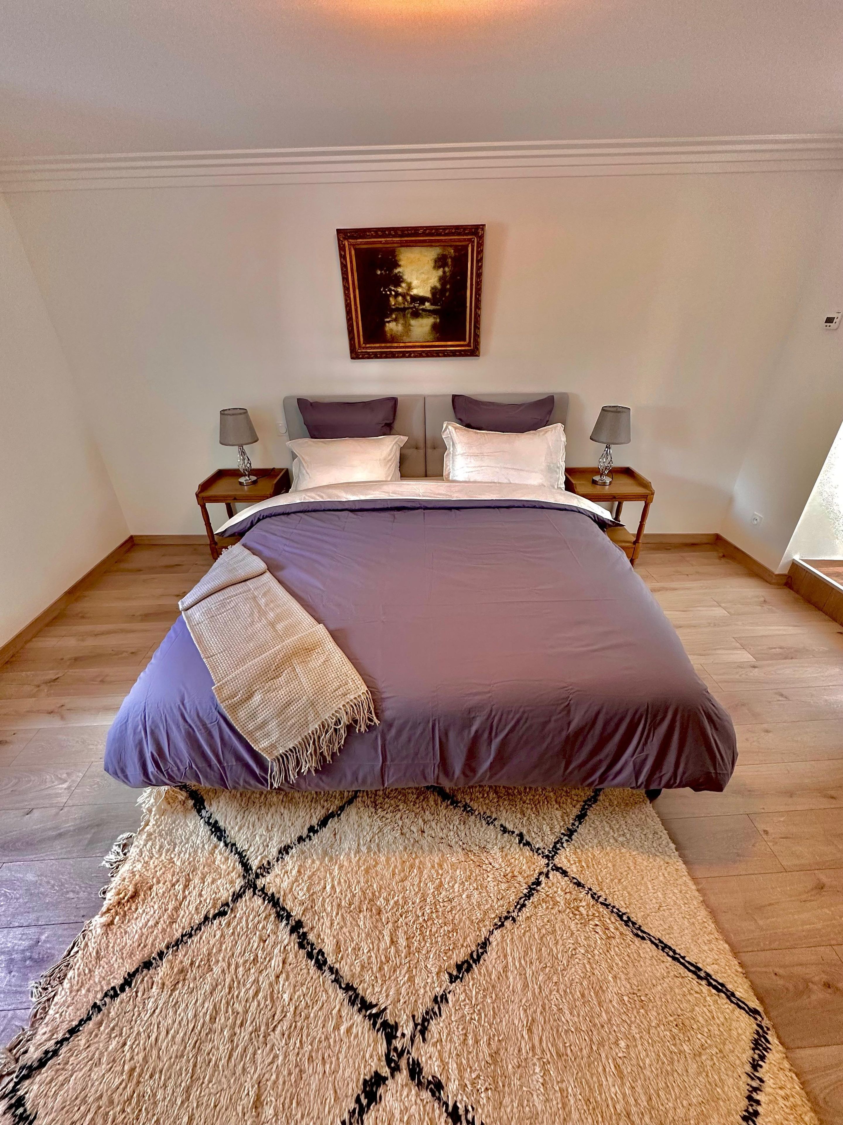 Un lit bien fait, avec une tête de lit, deux tables de chevet avec des lampes, un tableau accroché au mur et un tapis à motifs sur le sol.