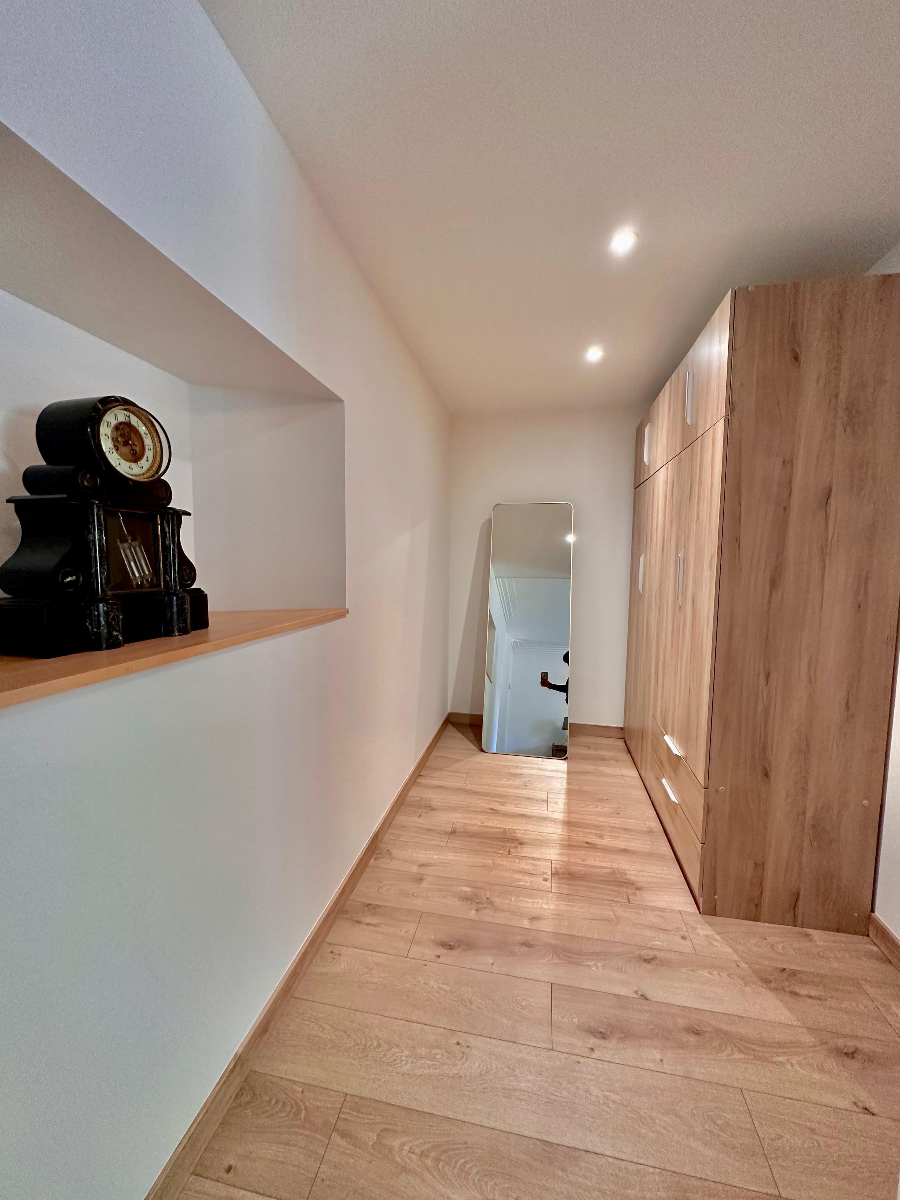 Un couloir doté d’un plancher en bois, d’une armoire en bois et d’une étagère sur laquelle se trouve une horloge.