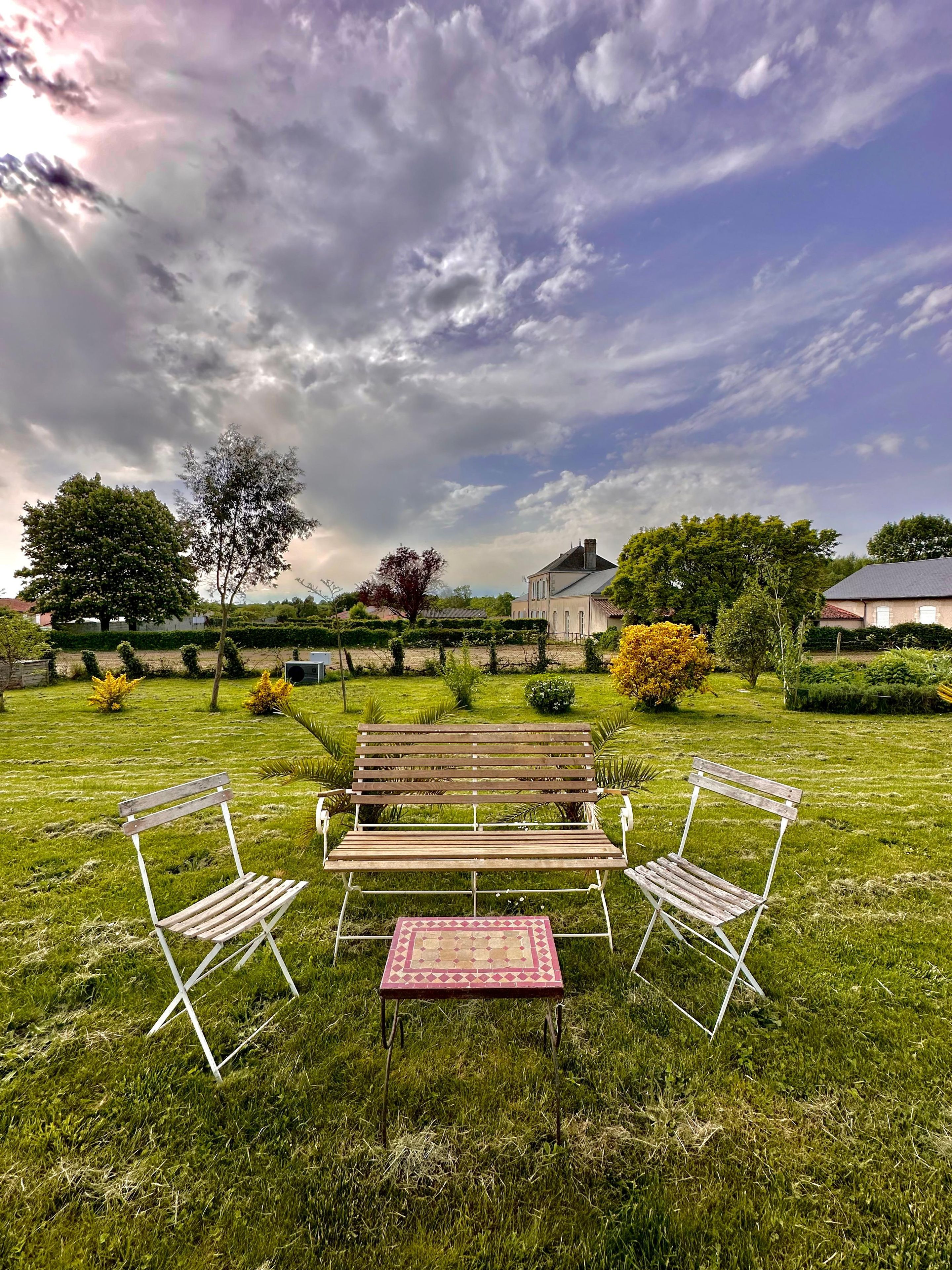 Un espace extérieur aménagé avec un banc en bois et deux chaises en métal, une petite table recouverte d’une nappe à motifs, et un champ verdoyant avec des arbres et une maison en arrière-plan.