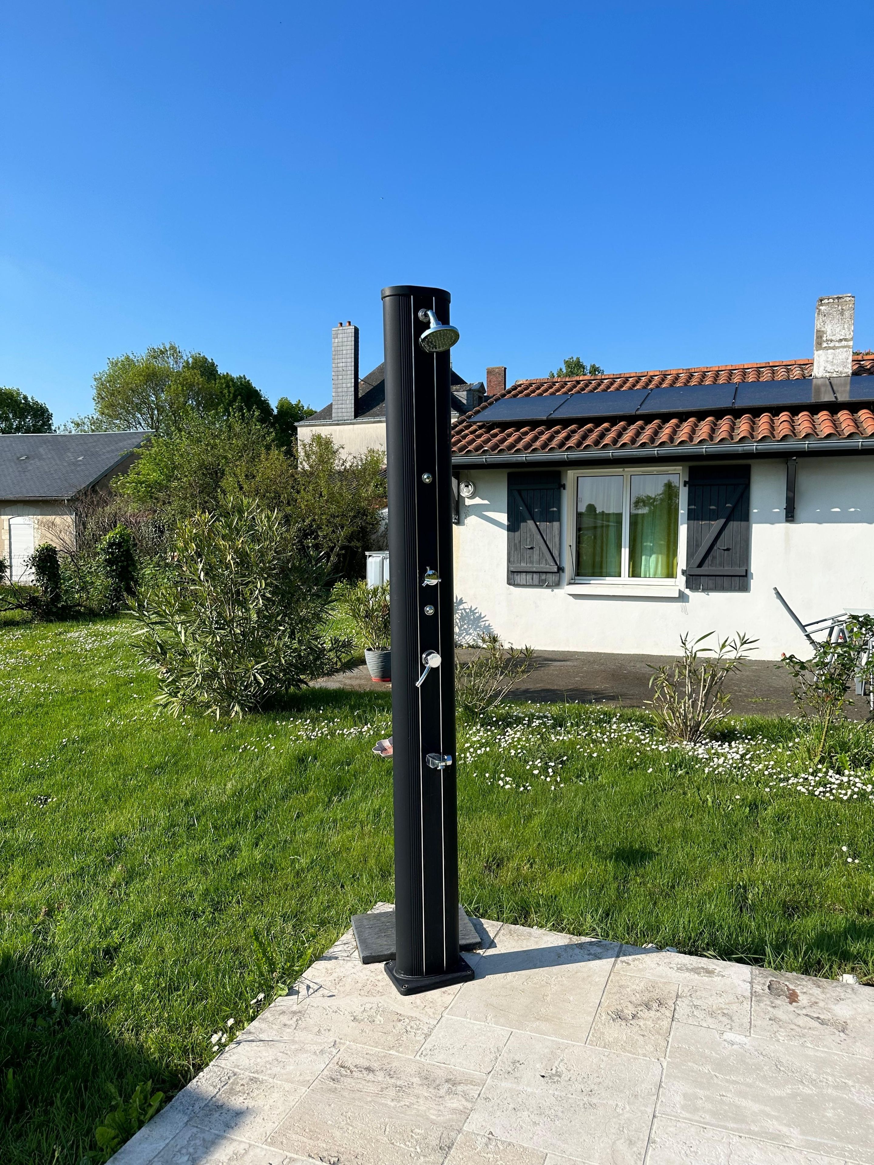 Une douche extérieure noire, dotée d’un pommeau de pluie, installée sur une plateforme en pierre dans un espace verdoyant, avec une maison en arrière-plan.
