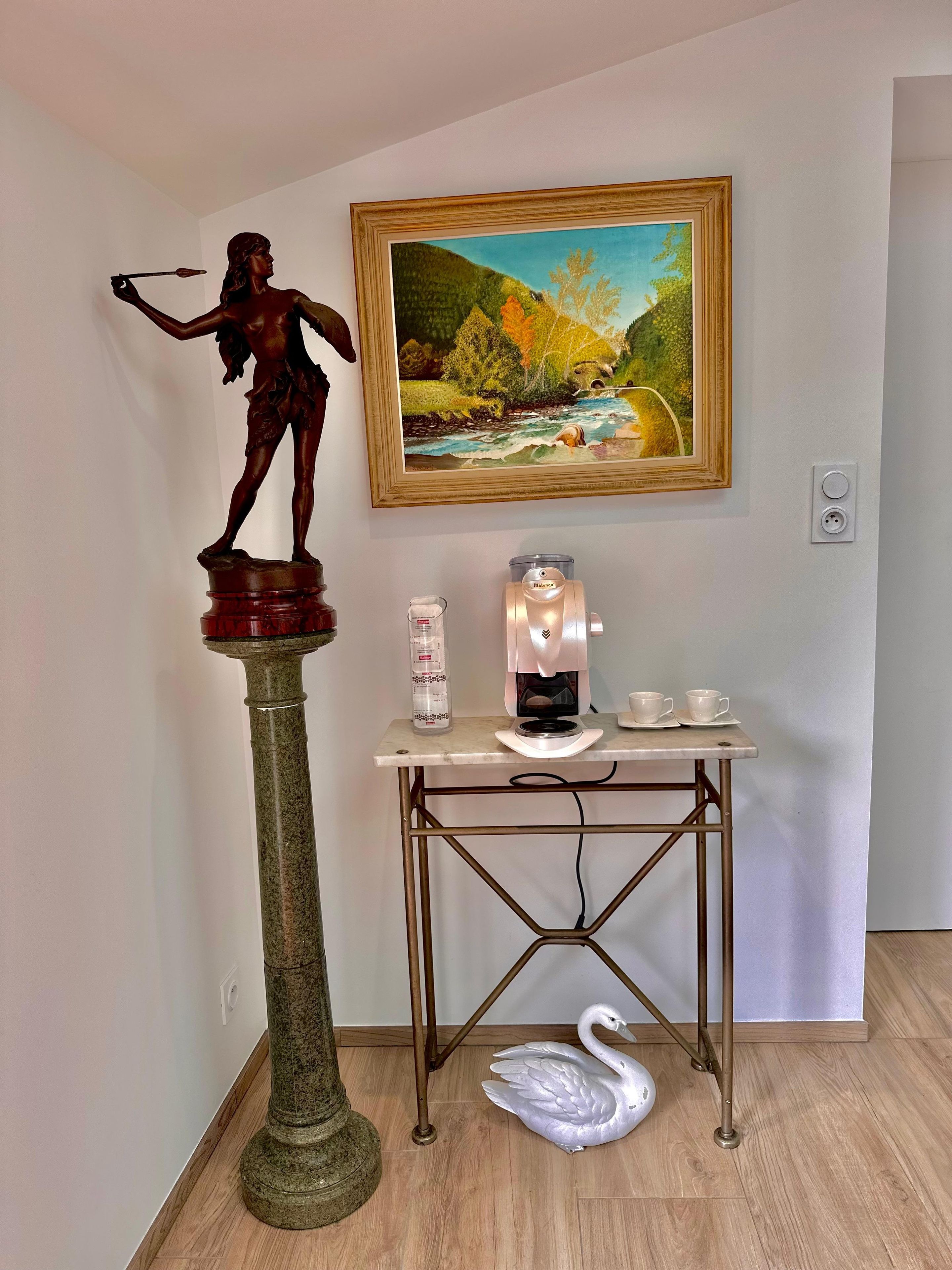Eine Statue auf einem Sockel, ein Gemälde an der Wand, eine Kaffeemaschine, Tassen und eine Schwaneinskulptur auf dem Boden.
