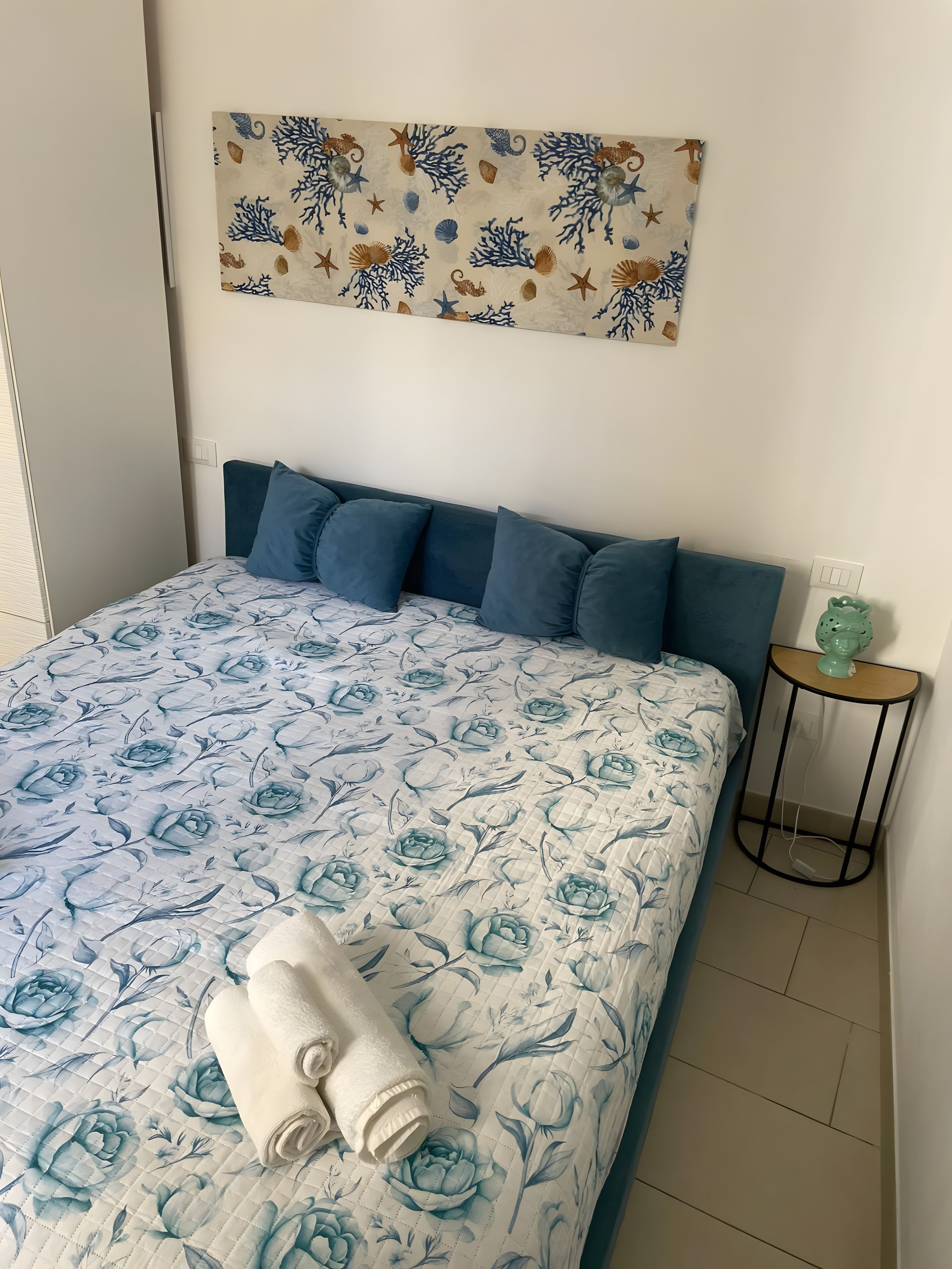Un letto con una coperta a fiori, una testiera con molti cuscini, un'opera d'arte appesa al muro e un tavolino con un oggetto verde sopra.