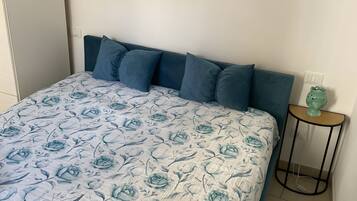 Un letto con una coperta a fiori, una testiera con molti cuscini, un'opera d'arte appesa al muro e un tavolino con un oggetto verde sopra.