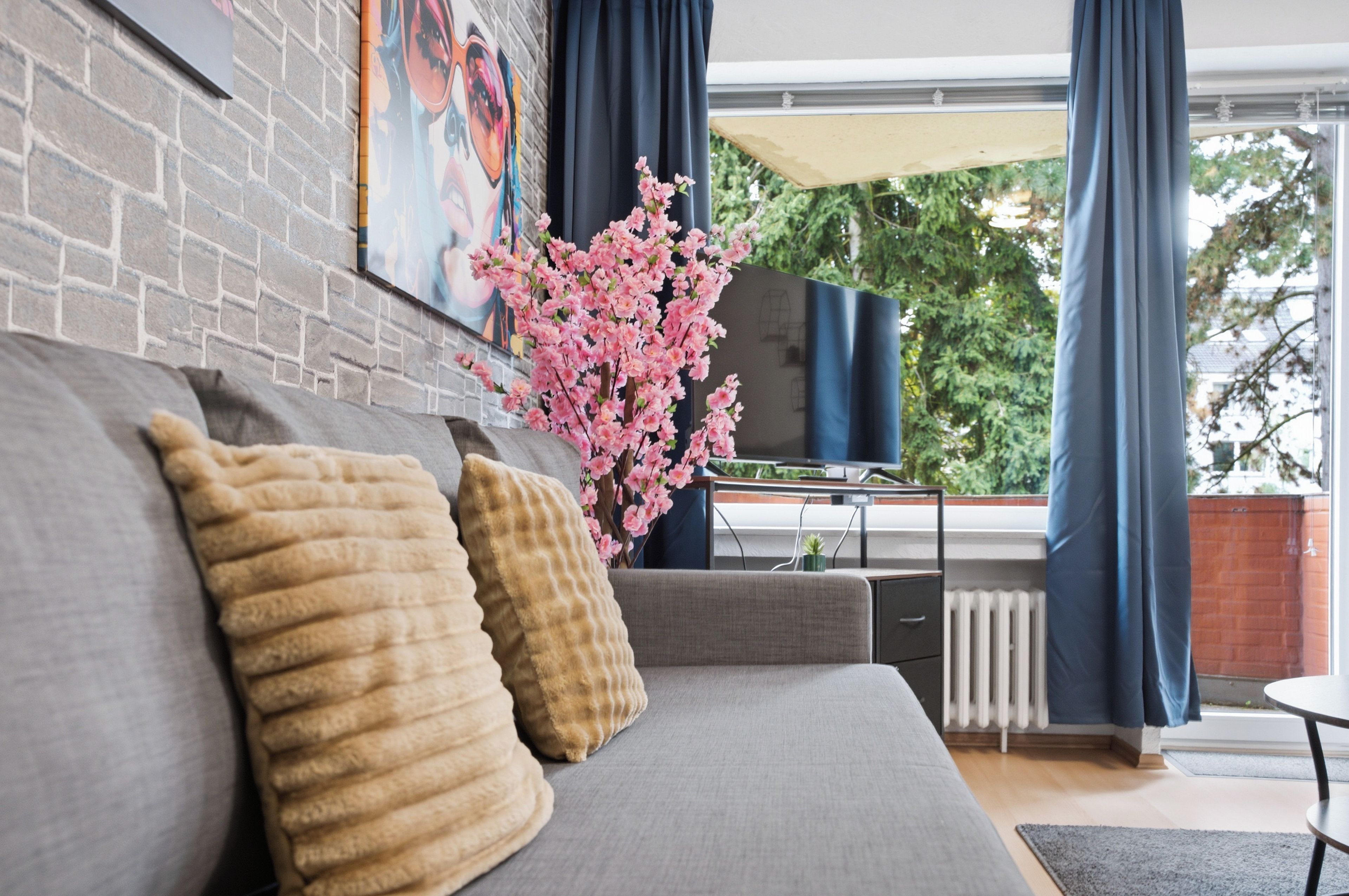 Ein modernes Wohnzimmer mit einem grauen Sofa, dekorativen Kissen, einer blühenden rosa Pflanze, einem Flachbildfernseher und einem Fenster mit Vorhängen.