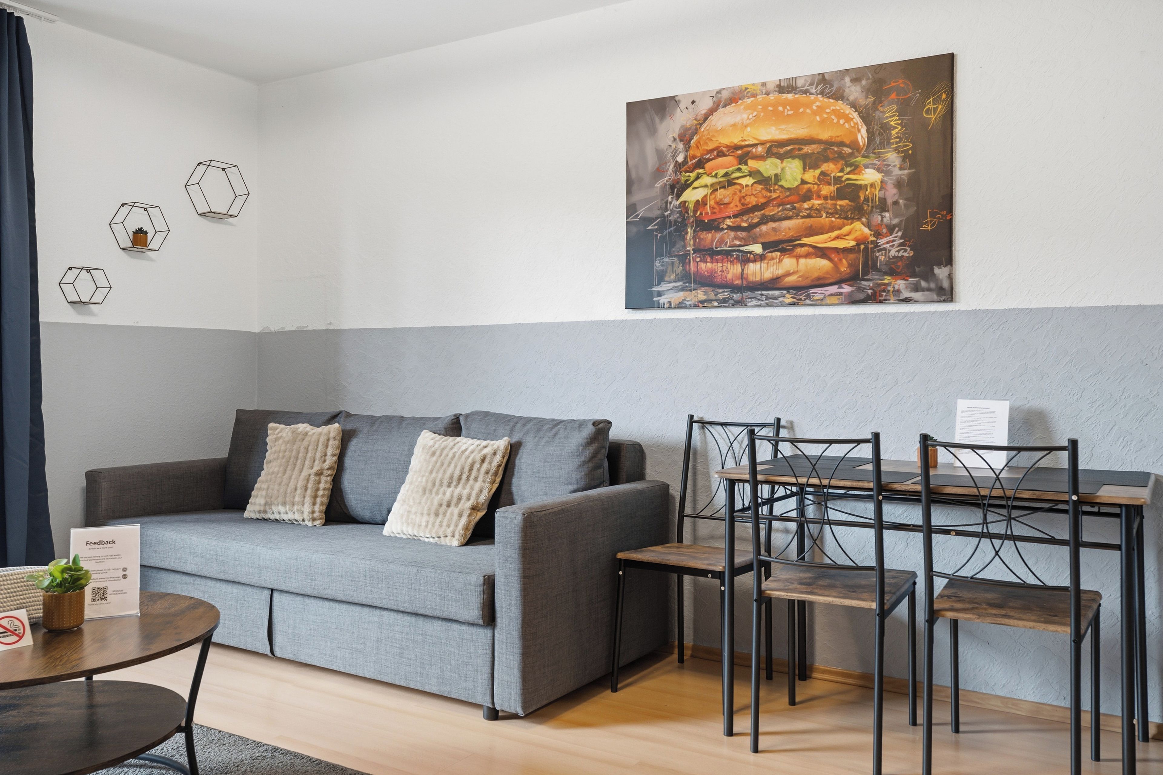 Ein modernes Wohnzimmer mit einem grauen Sofa, einem Esstisch mit Stühlen und einem großen Gemälde eines Burgers an der Wand.