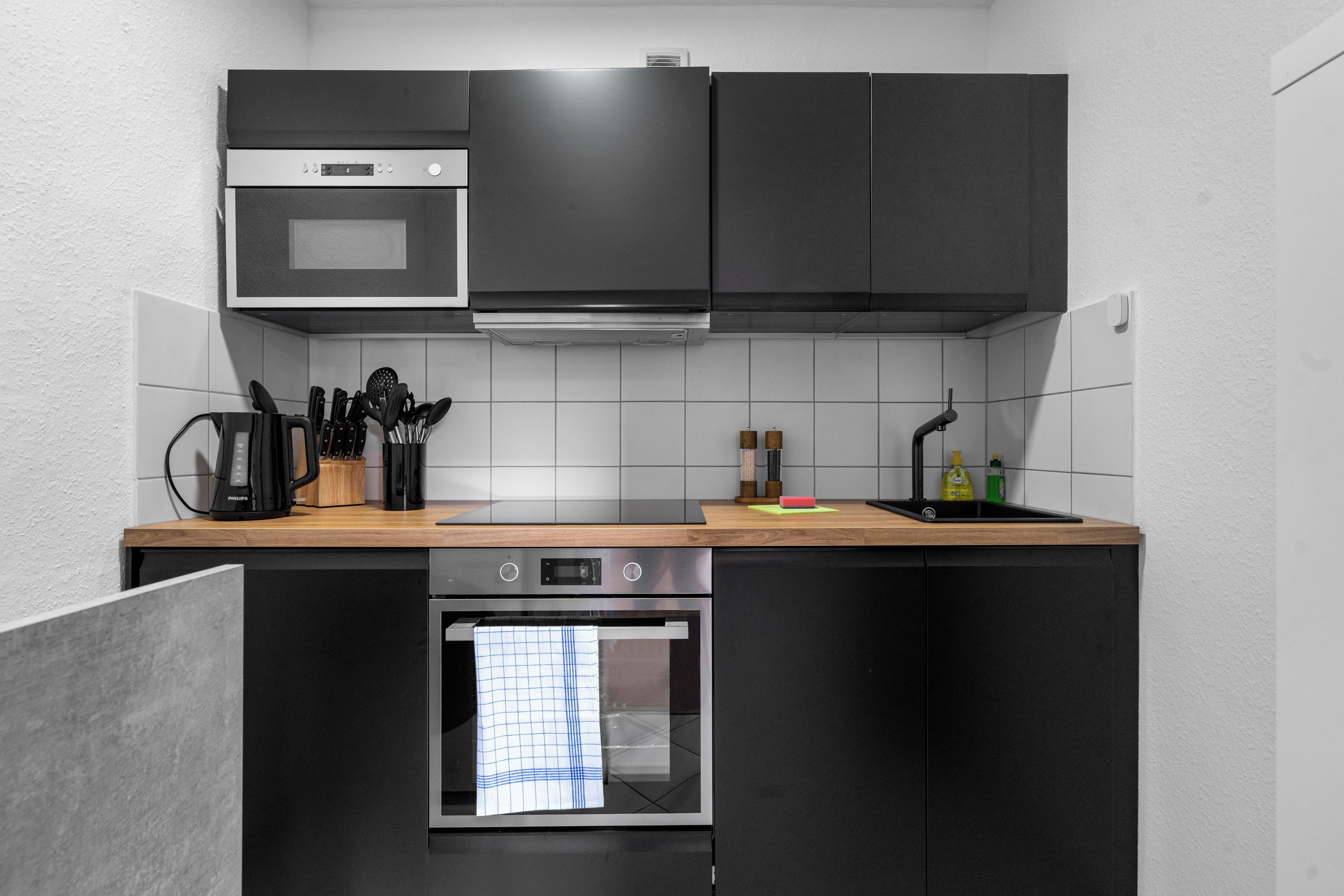 Une cuisine moderne avec des meubles de couleur noire, un plan de travail en bois, un four encastré et un four à micro-ondes.