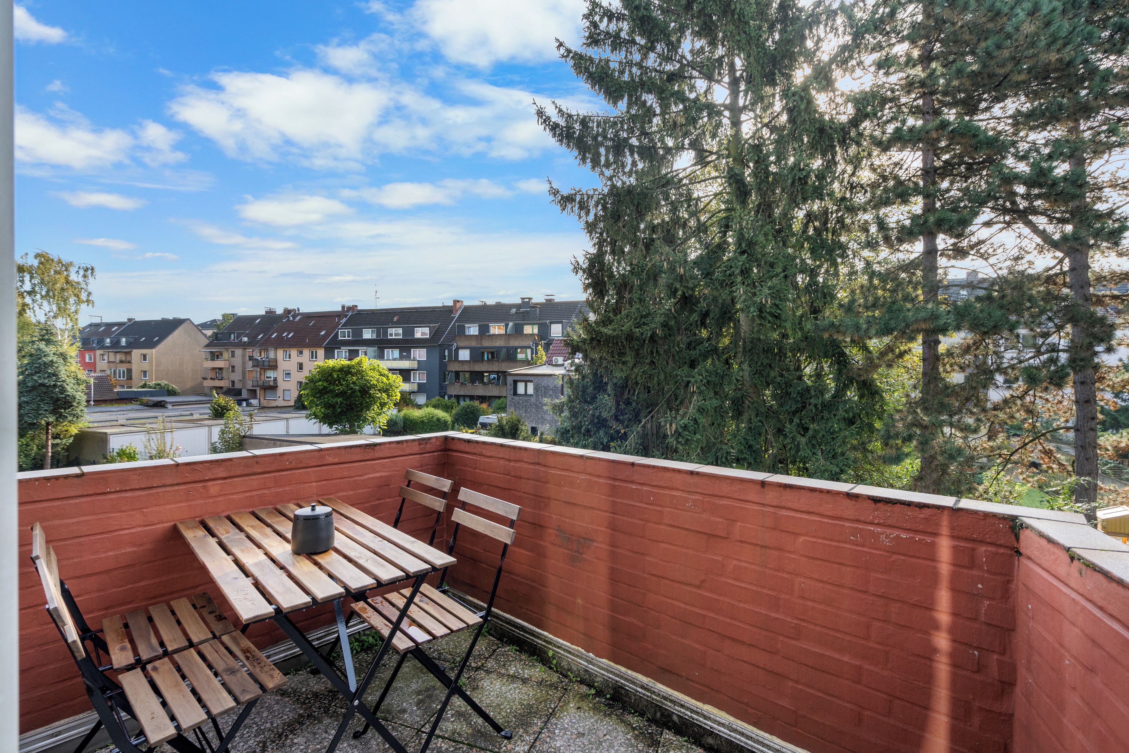 Un balcon avec une table et des chaises en bois, un mur en briques rouges et une vue sur des immeubles résidentiels et des arbres.