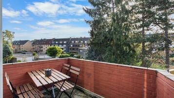 Un balcon avec une table et des chaises en bois, un mur en briques rouges et une vue sur des immeubles résidentiels et des arbres.