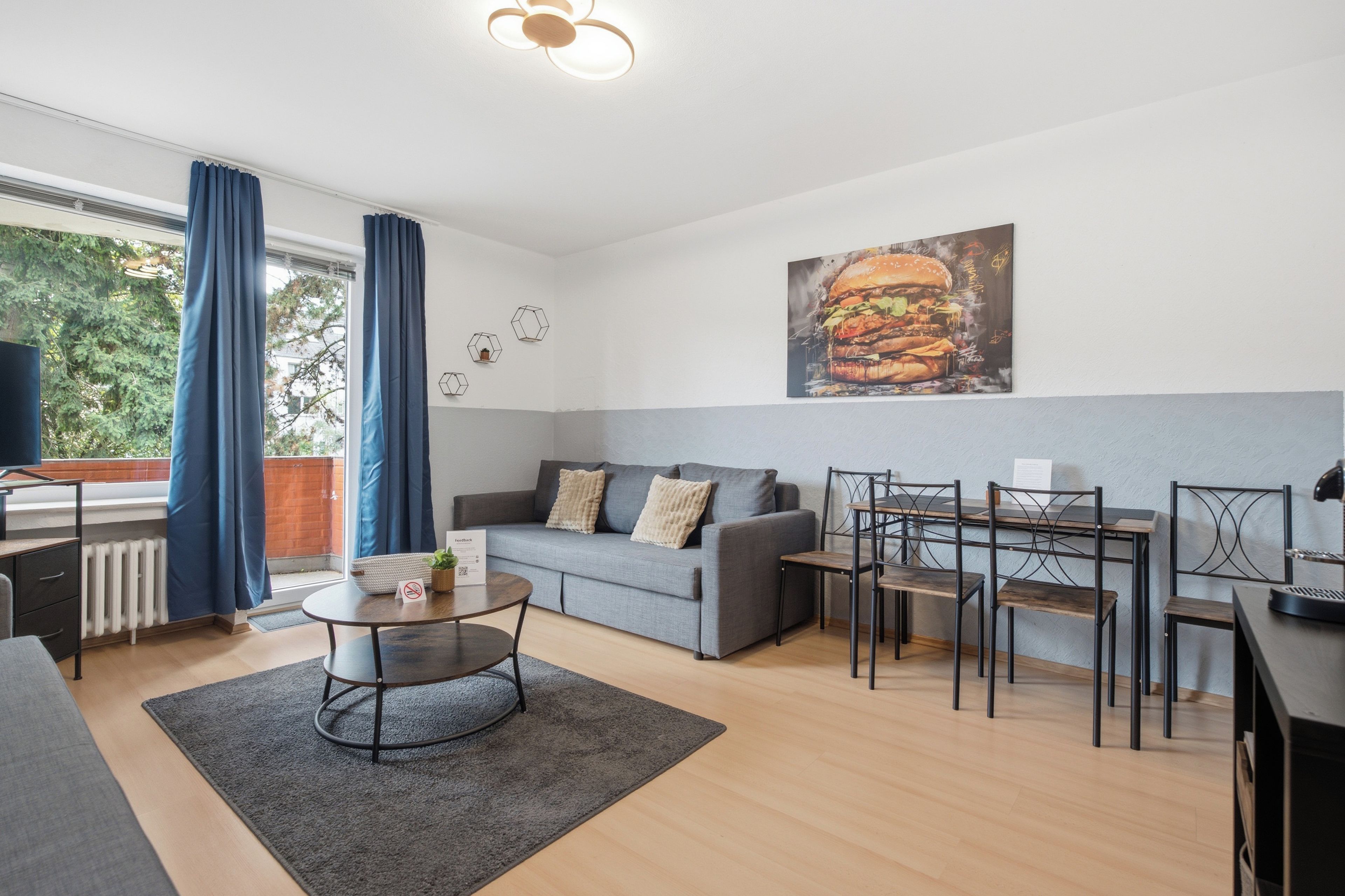 A modern living room with a grey sofa, a dining table with chairs, a coffee table, and a large painting of a burger.