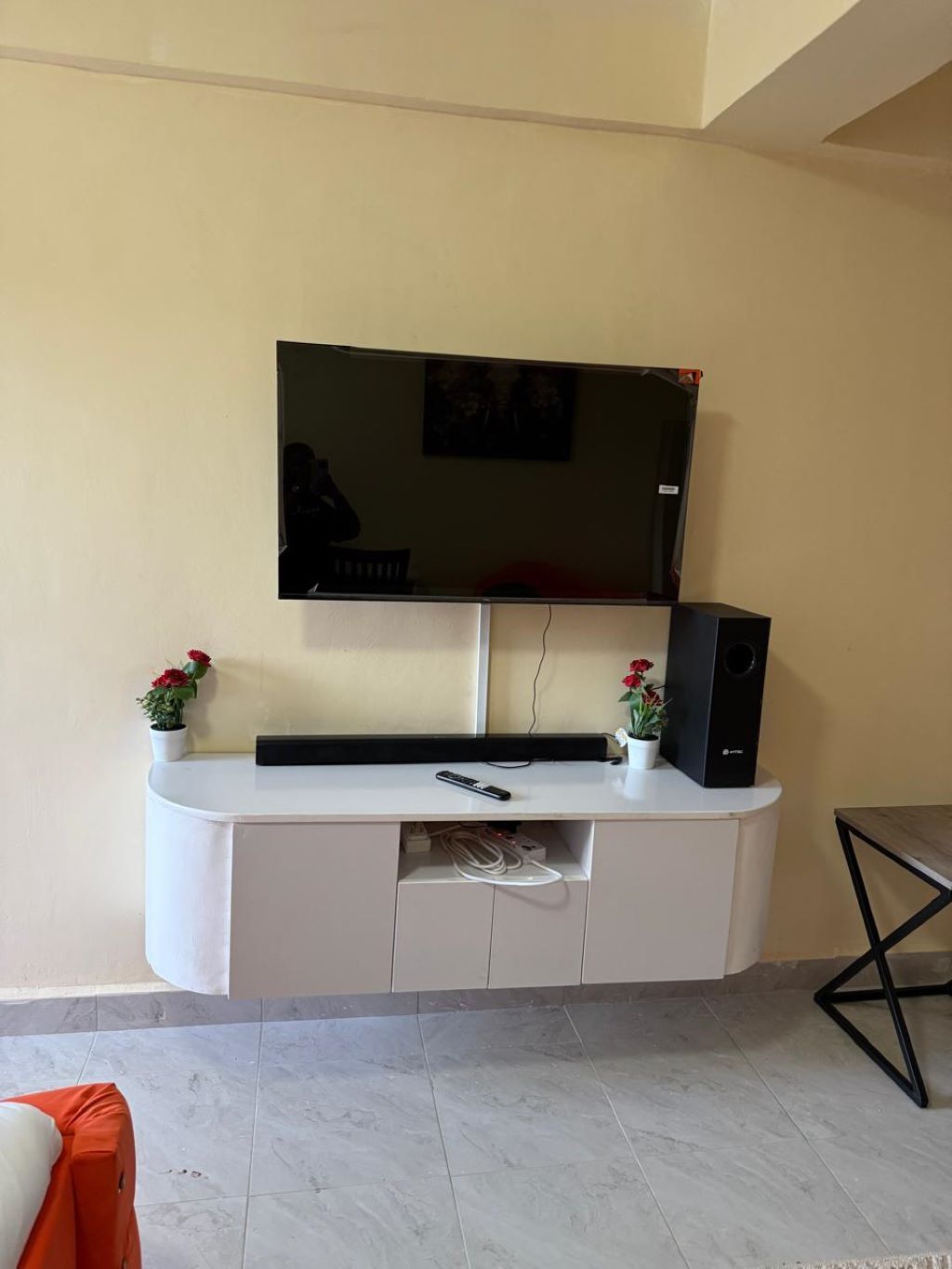 Un televisor de pantalla plana montado en la pared sobre un mueble blanco con un altavoz y un control remoto.