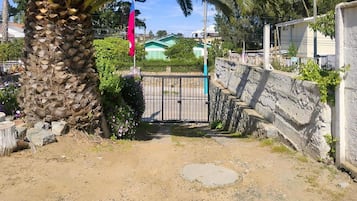 A gated entrance with a palm tree and a flag, leading to a residential area with houses and a green building in the background.