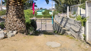 A gated entrance with a palm tree and a flag, leading to a residential area with houses and a green building in the background.
