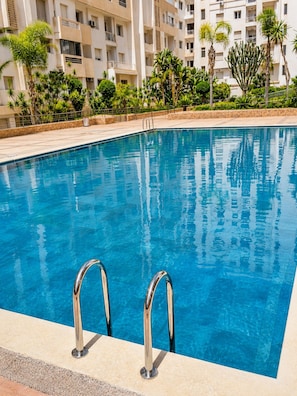 A swimming pool with metal handrails, surrounded by a landscaped area with palm trees and a multi-story building in the background. - New Listing (Agadir)