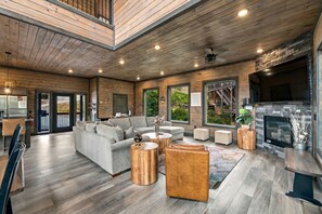 A spacious living area with a fireplace, wooden flooring, and a large window overlooking greenery. - Modern 4BR Cabin within minutes of Gatlinburg. Hot Tub! (Gatlinburg)