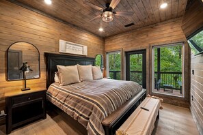 Ein Schlafzimmer mit Holzbalkendecke, einem Bett mit gestreifter Bettwäsche, einem Nachttisch mit Lampe, einem Spiegel und einem Fenster mit Blick auf Bäume.