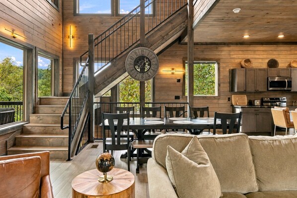 A spacious interior with a wooden staircase, a dining area with a round table, and a kitchen with modern appliances. - Modern 4BR Cabin within minutes of Gatlinburg. Hot Tub! (Gatlinburg)