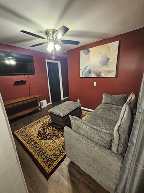 Una sala de estar con ventilador de techo, televisor de pantalla plana, sofá, mesa de centro, alfombra estampada y una puerta.