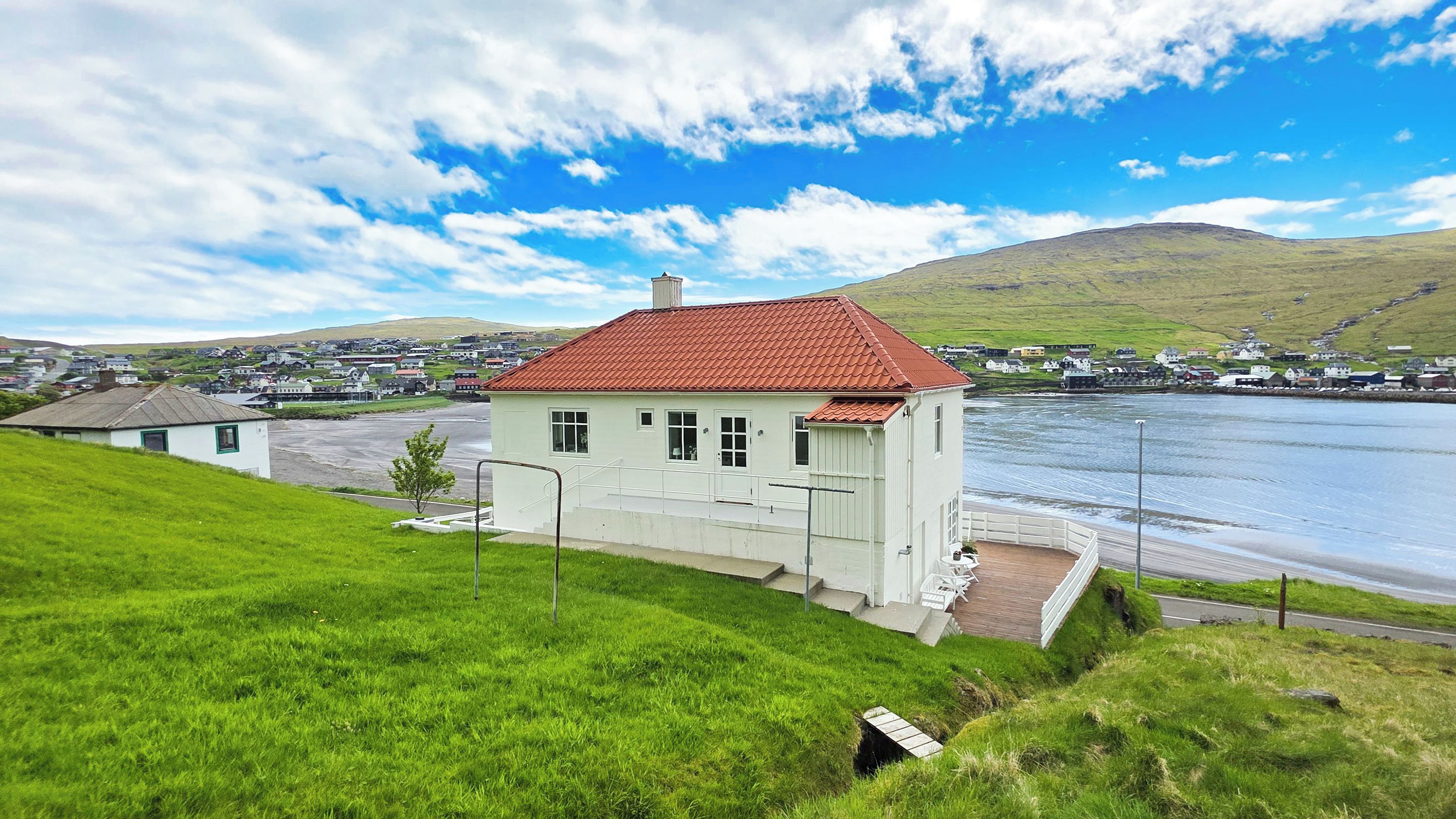 A coastal building with a red roof, a small porch, and a view of the sea and a town in the background.