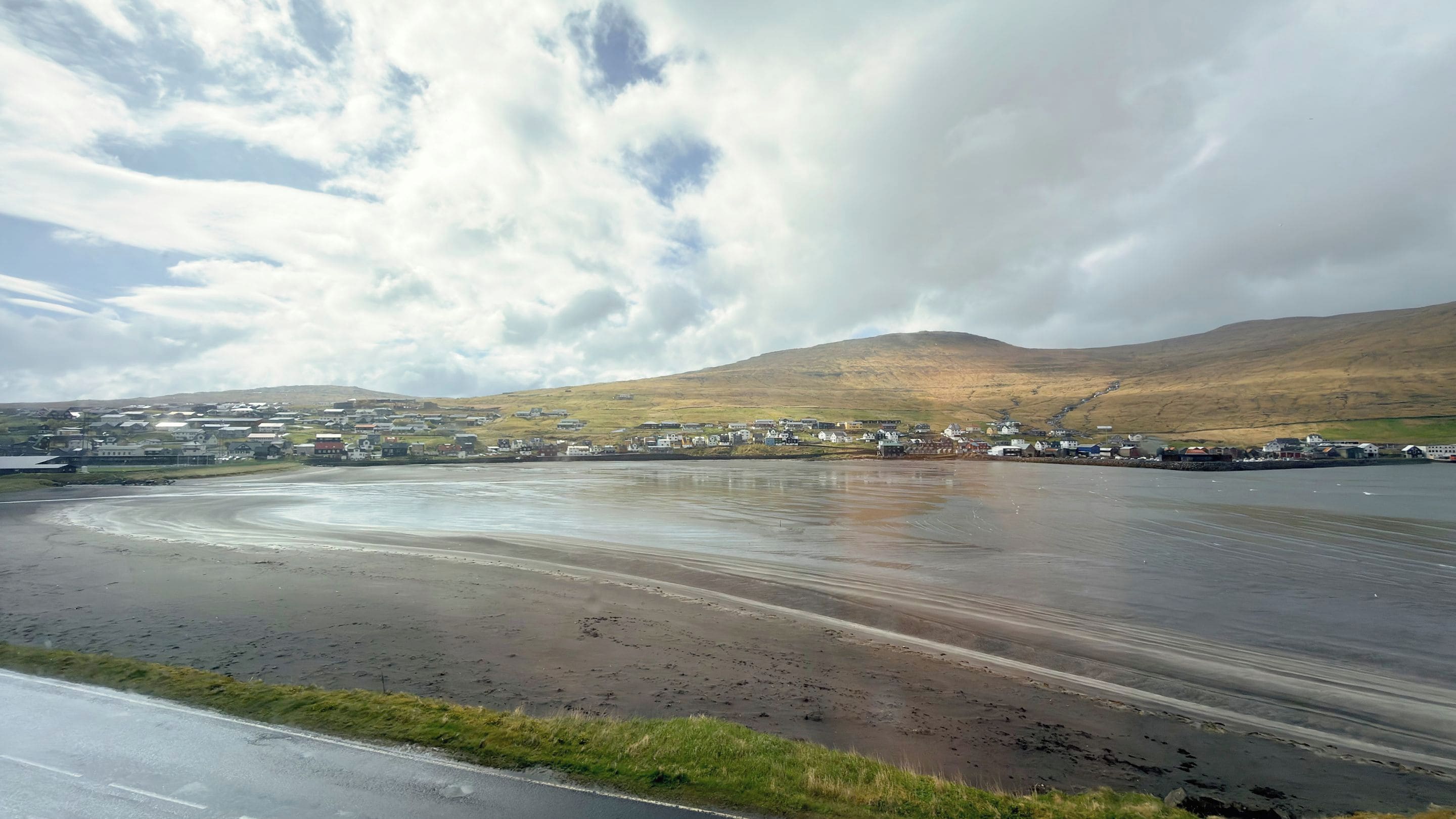 A coastal road with a wet surface, a grassy verge, and a town in the background with buildings and hills.
