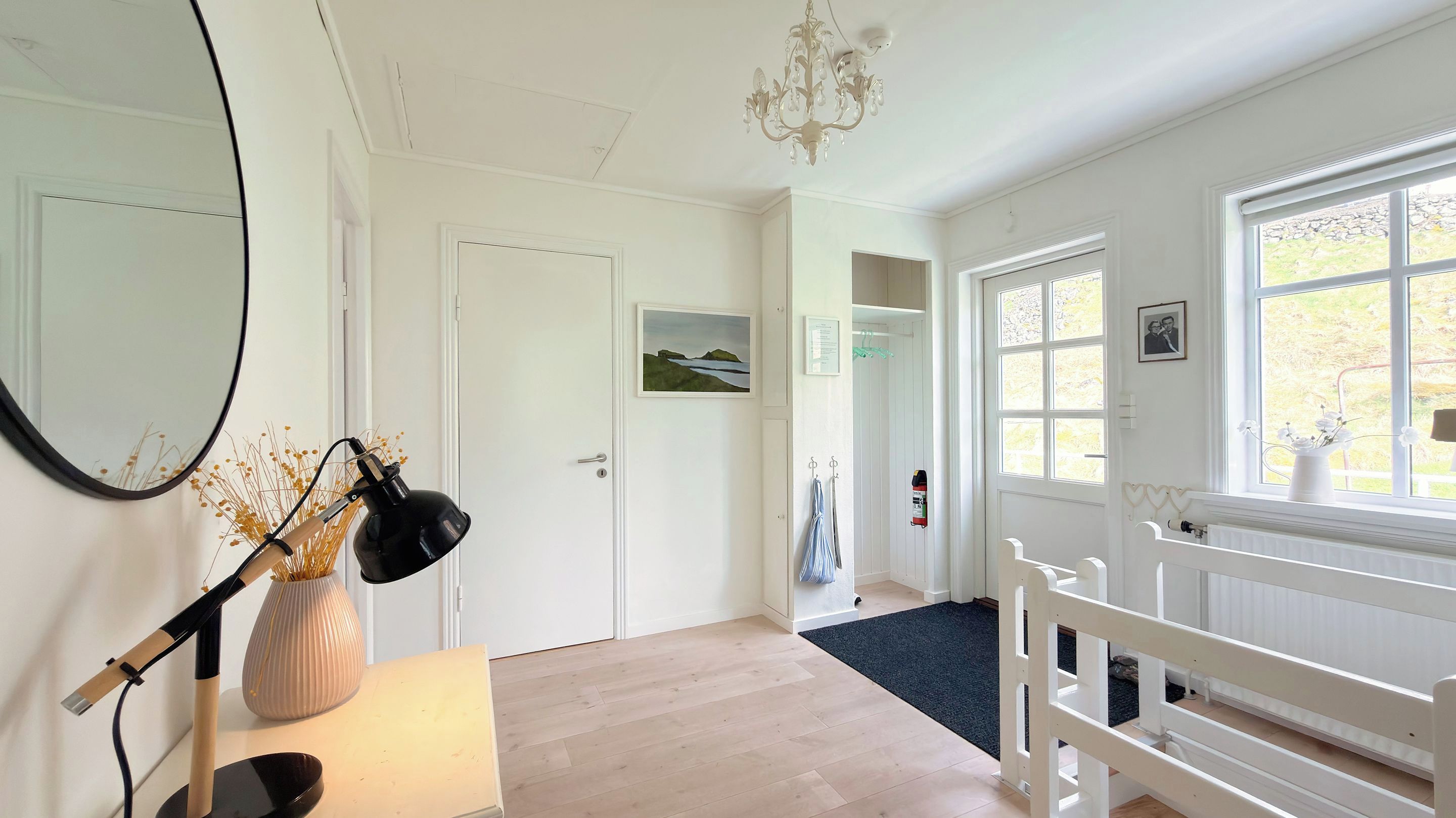A room with a wooden floor, white walls, a chandelier, a large window, and a door with a painting.